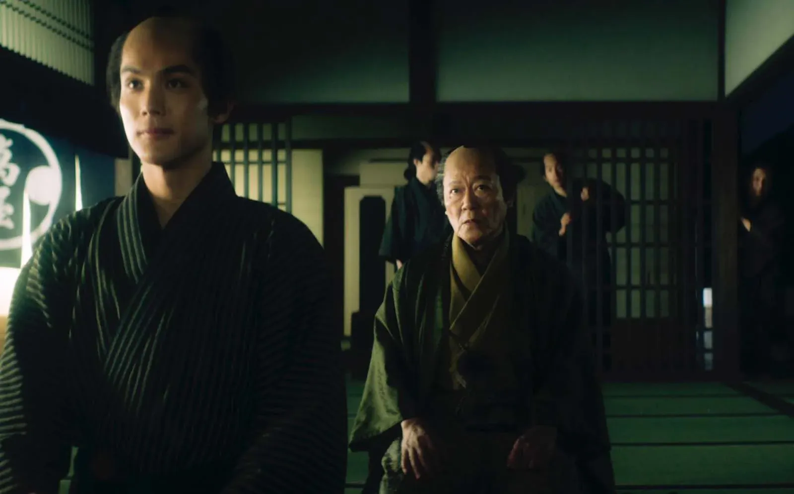 Un homme en kimono au premier plan, devant un homme âgé assis dans un intérieur japonais sombre.