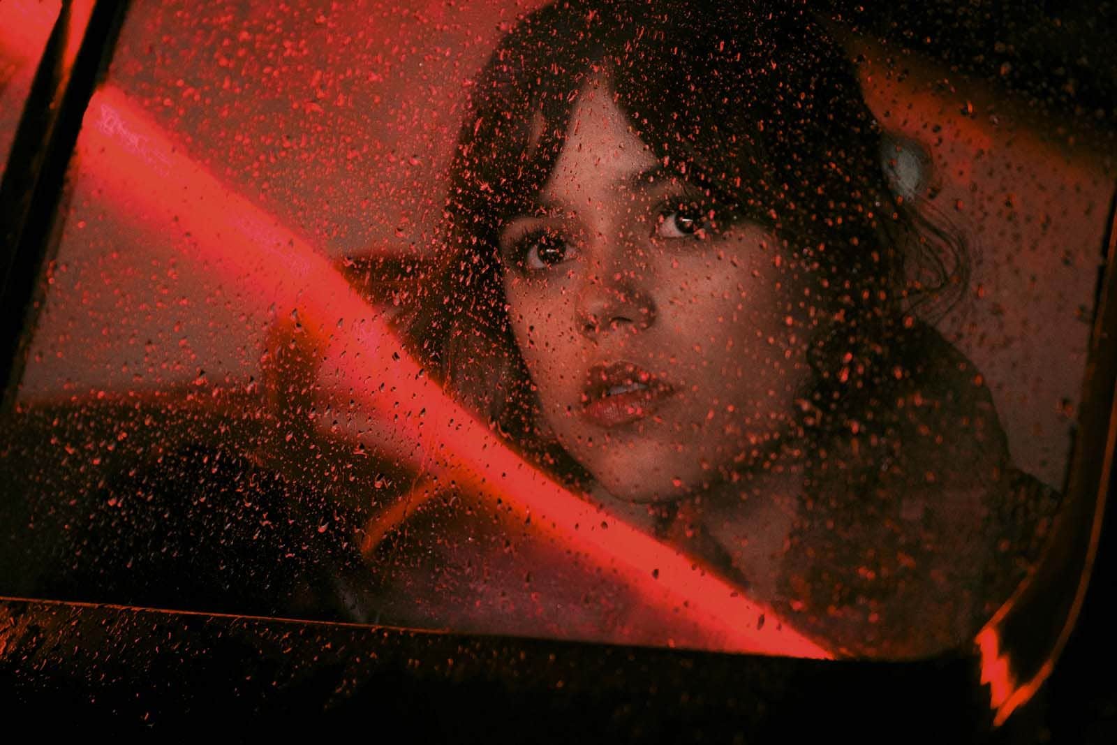 Femme regardant à travers une vitre mouillée, éclairée par une lumière rouge à l’intérieur d’une voiture