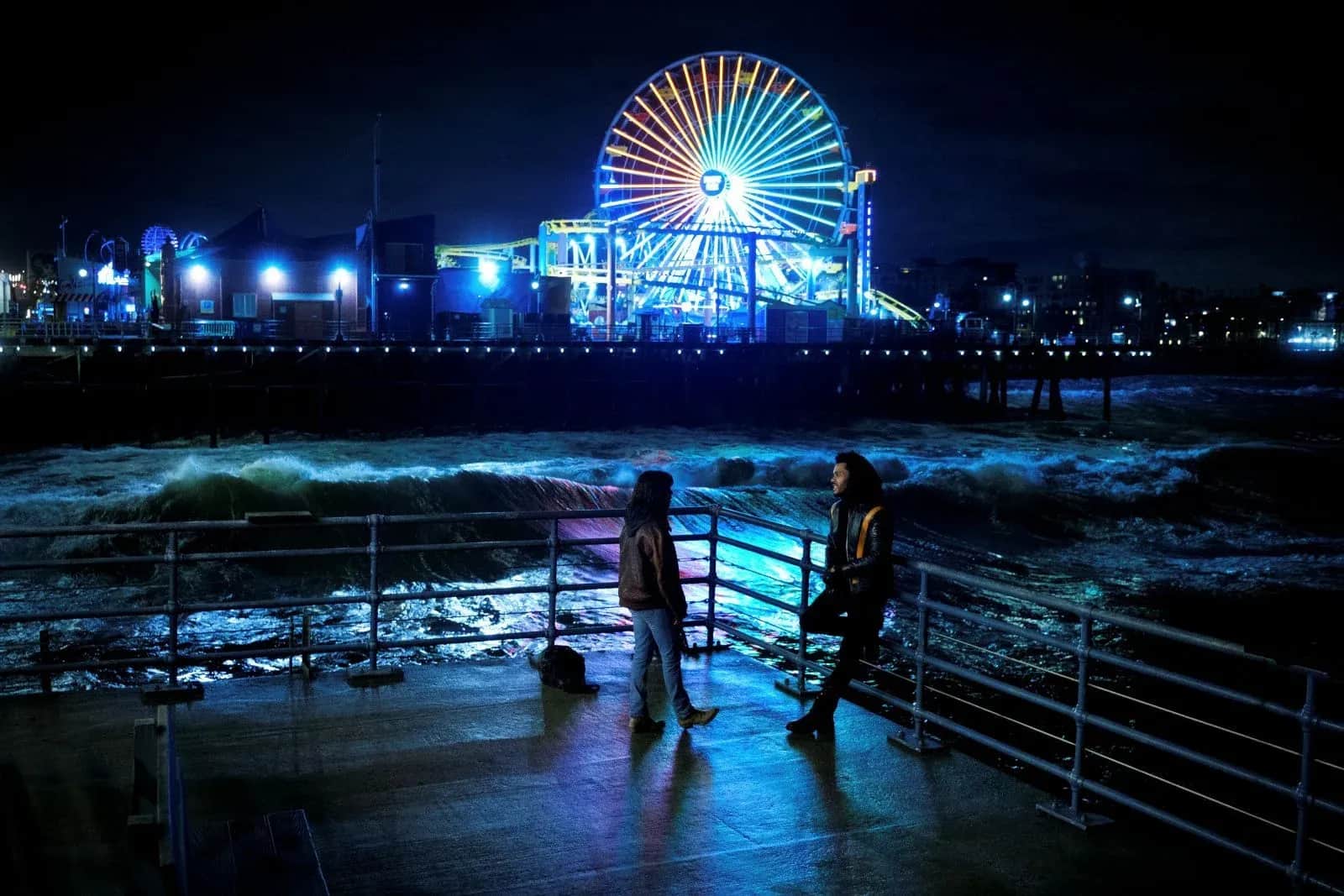 Deux personnes se tiennent sur une jetée face aux vagues, avec une grande roue illuminée en arrière-plan.