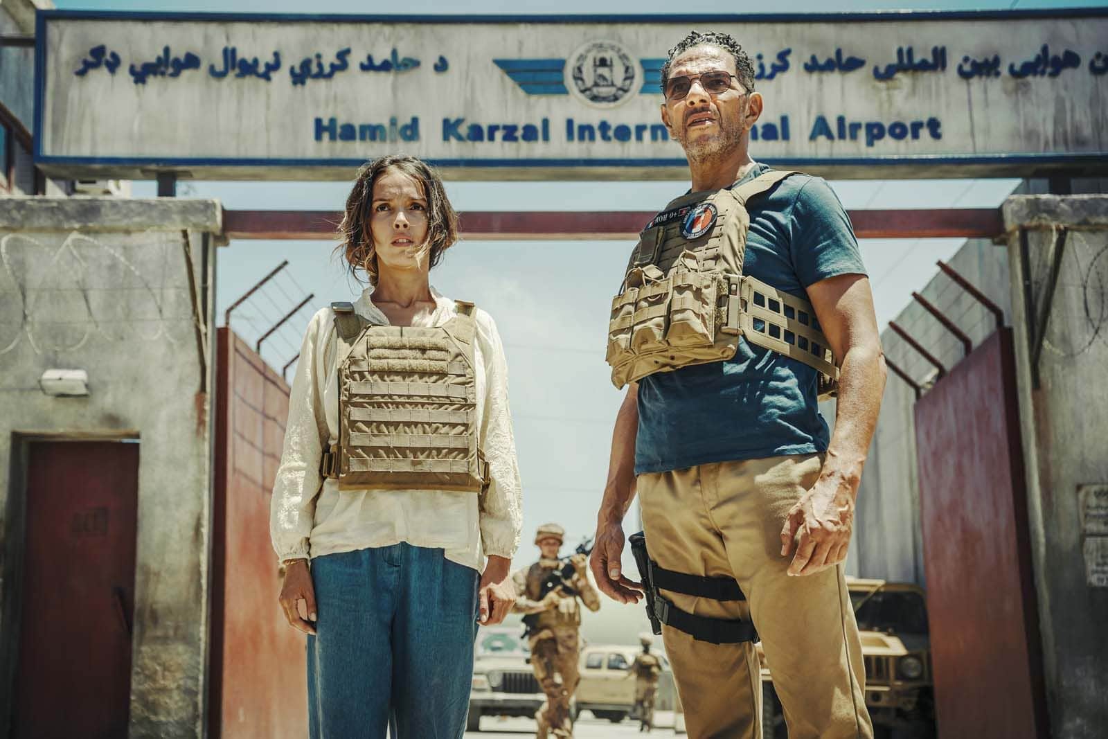 Une femme et un homme en gilets pare-balles devant l’aéroport international Hamid Karzai, avec des soldats en arrière-plan.