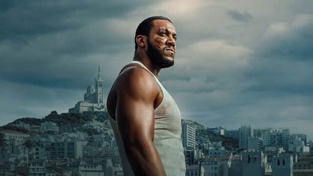 Un homme musclé en débardeur blanc regarde au loin, avec Marseille en arrière-plan sous un ciel nuageux.