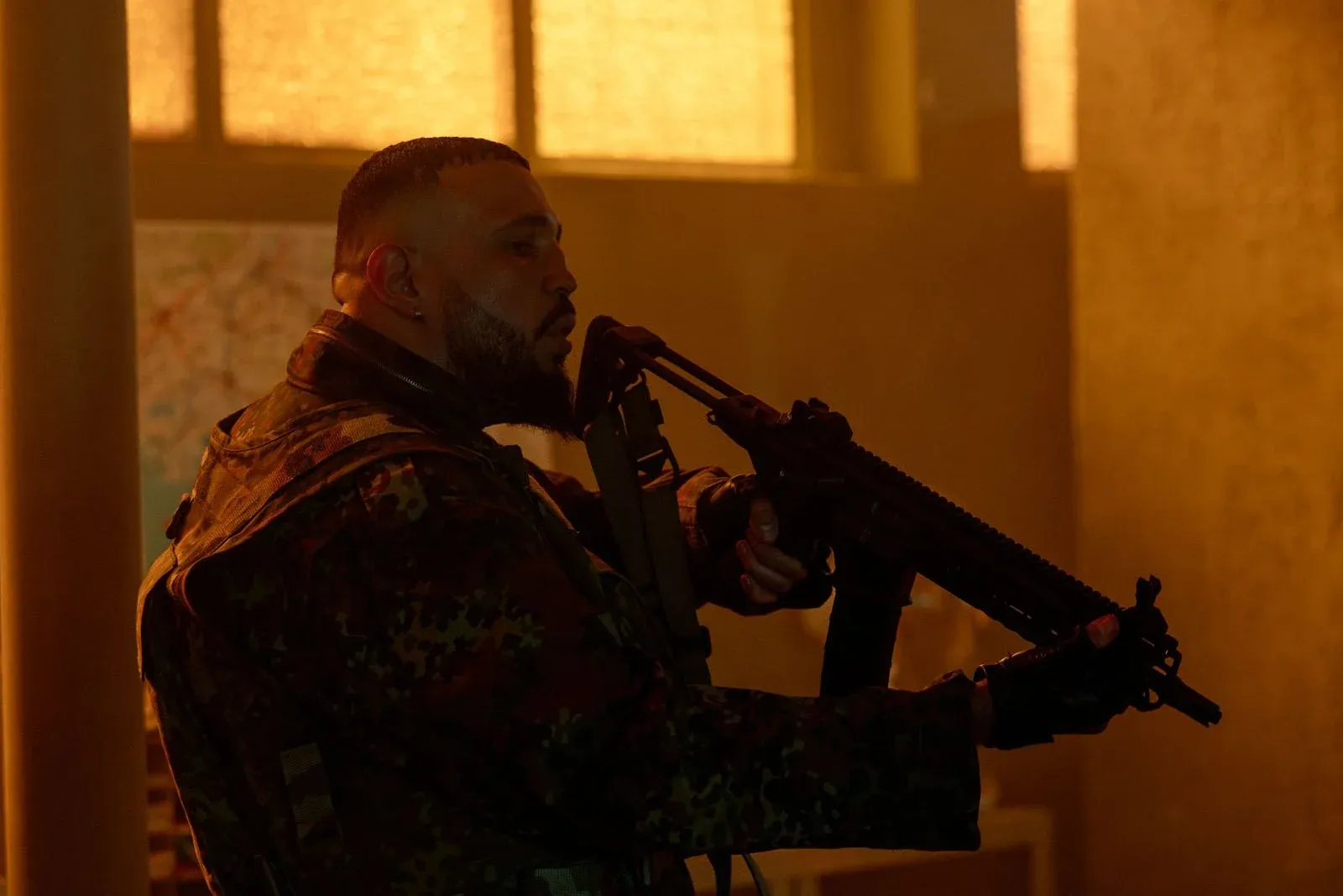 Un homme barbu en tenue militaire tient un fusil dans un bâtiment baigné de lumière orange.