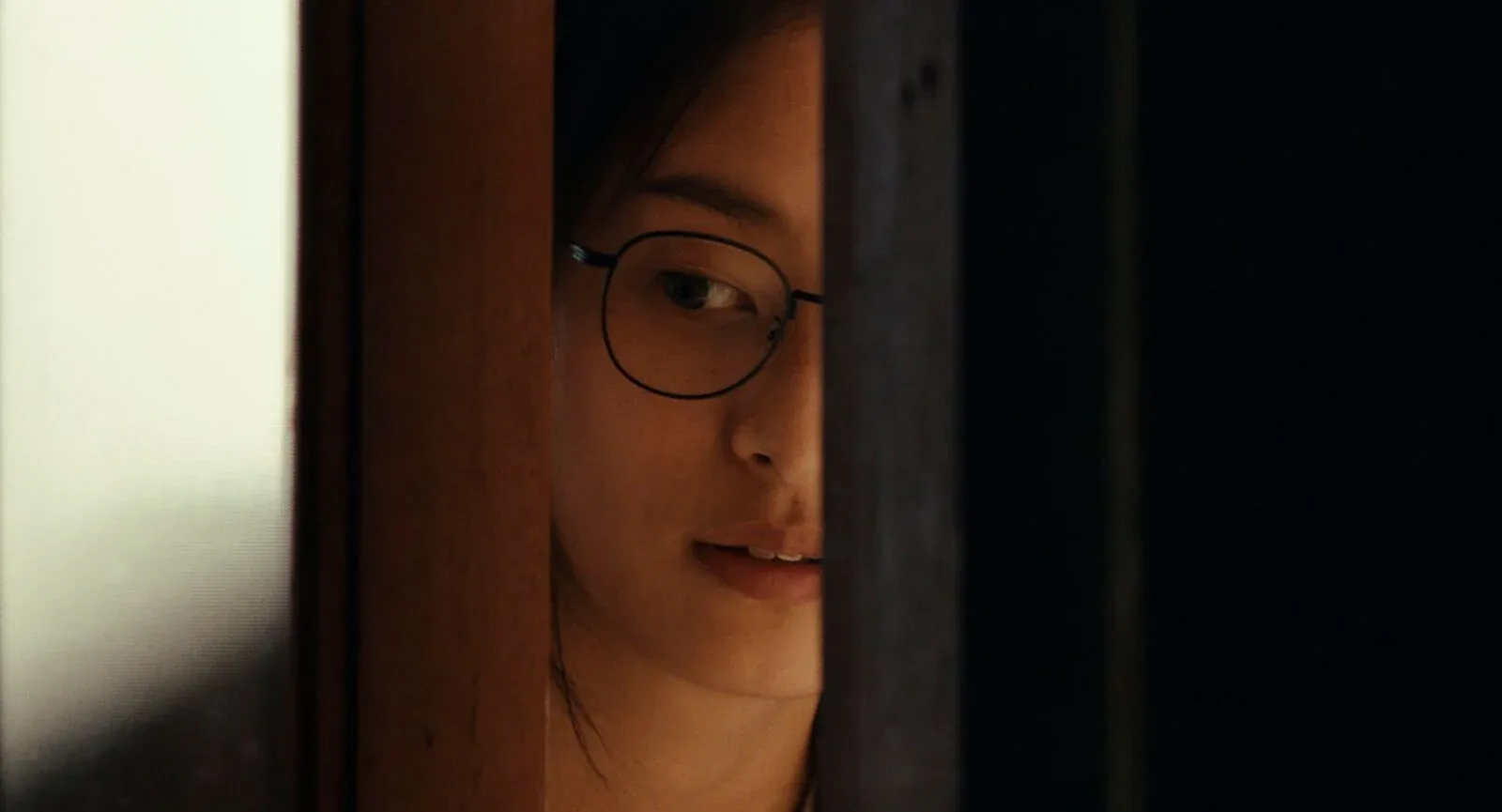 Une femme portant des lunettes observe à travers l’entrebâillement d’une porte.
