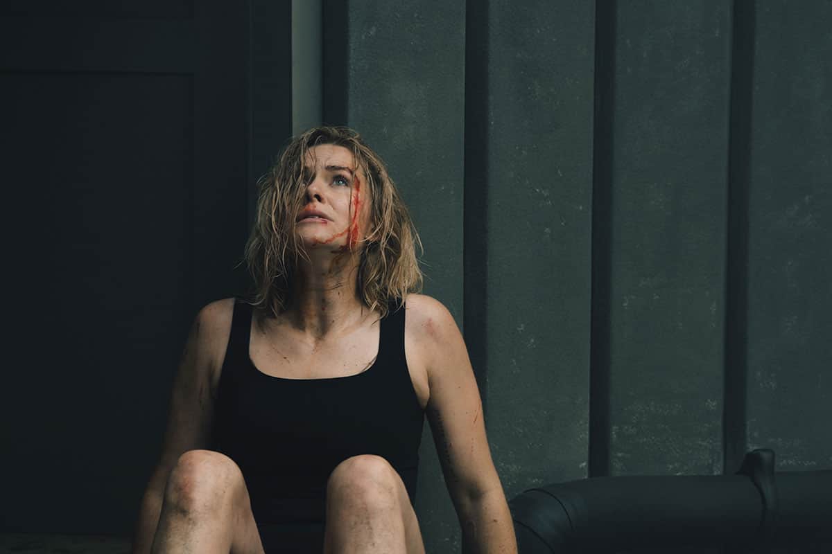 Femme blessée assise au sol, le visage ensanglanté, devant un mur sombre