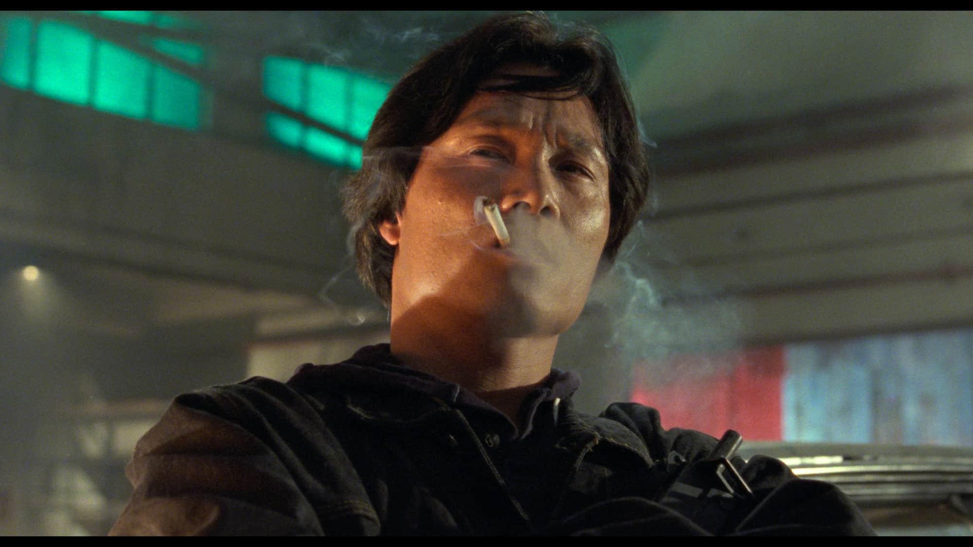Homme fumant une cigarette, avec de la fumée au visage, dans un intérieur éclairé de néons verts.