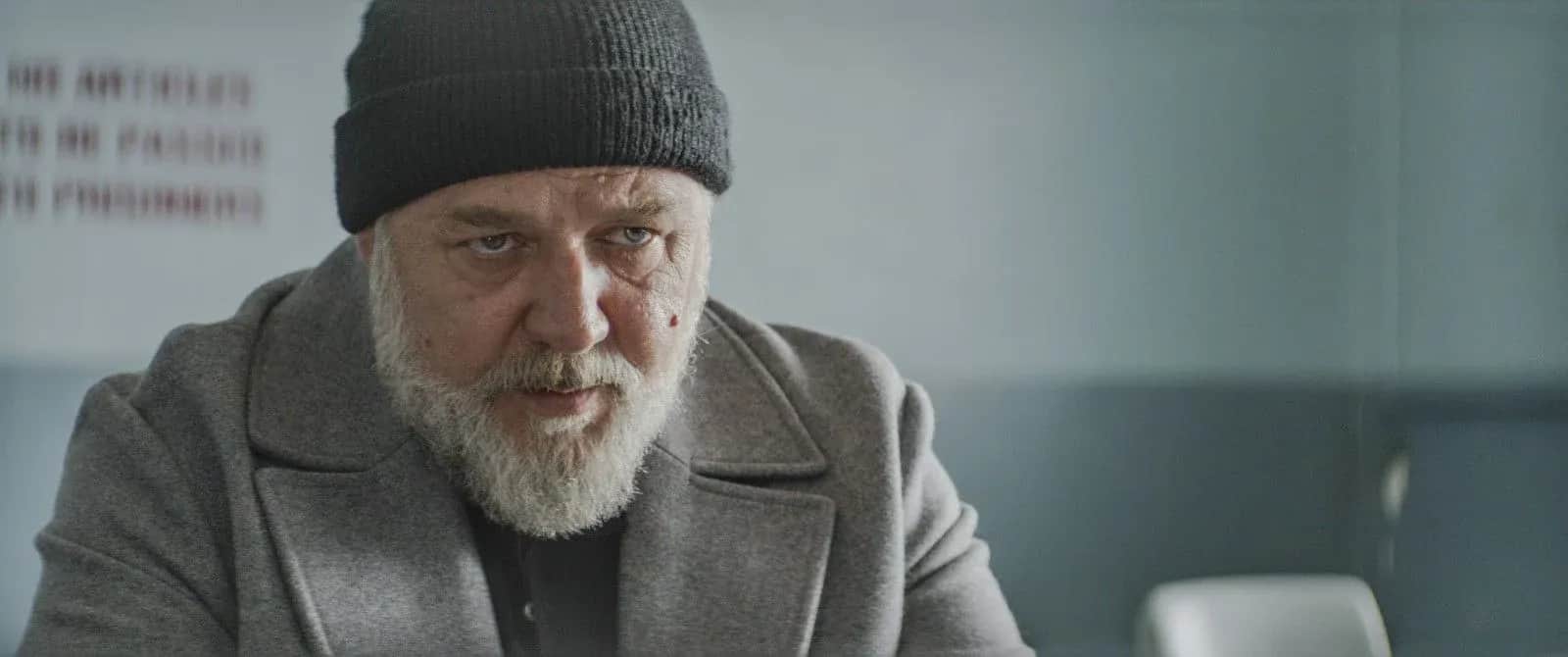 Homme barbu portant un bonnet et un manteau gris, assis dans une pièce au décor sobre