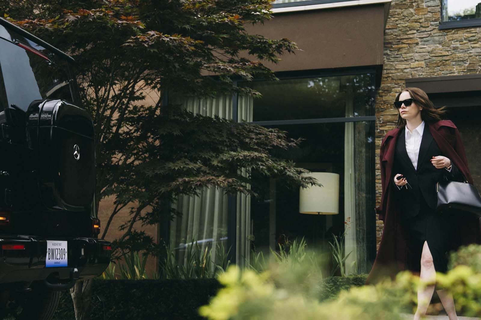Femme en lunettes noires sortant d’un véhicule noir devant une maison moderne, avec un sac à main.
