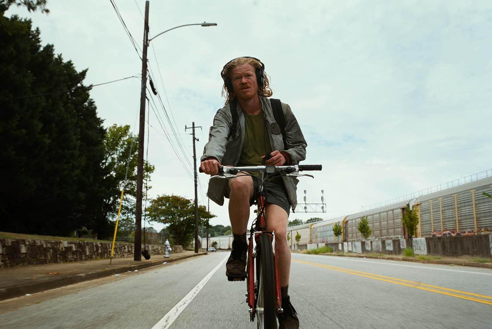 Homme à vélo portant un casque audio, roulant sur une route bordée d’arbres et de bâtiments industriels