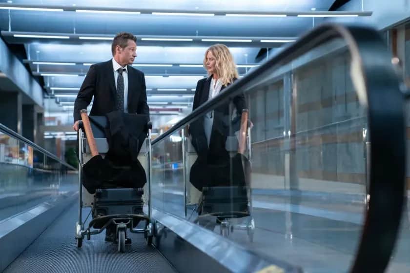 Un homme en costume pousse un chariot à bagages, accompagné d’une femme, dans un aéroport.
