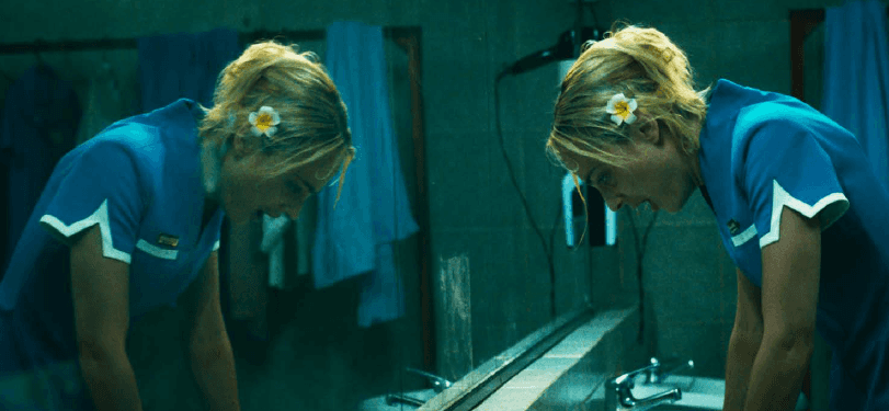 Une femme blonde en blouse bleue se penche au-dessus d’un lavabo, face à son reflet dans un miroir.